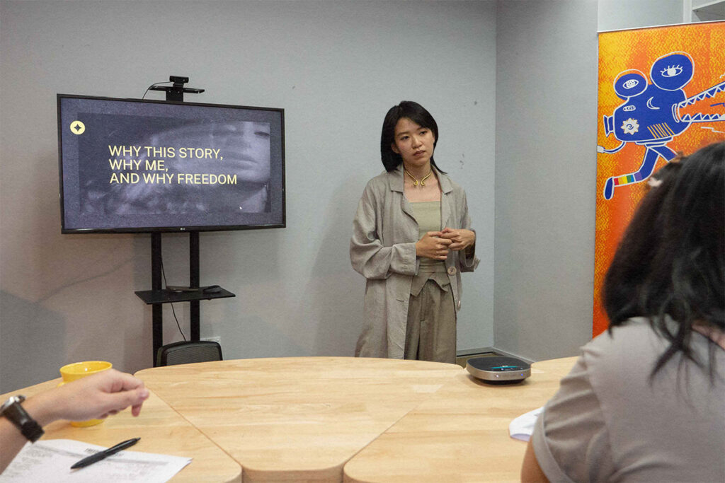 Zinc Chew during the Freedom Film Grant pitch 2023.  She is standing next to a TV screen where a page from her deck is visible and reads 'Why This Story, Why Me, And Why Freedom'